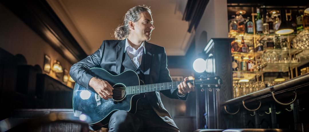 Ein Mann in einem Anzug spielt eine akustische Gitarre in einer schwach beleuchteten Bar, lächelt und schaut in die Kamera.