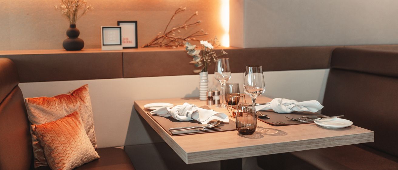 Ein elegant gedeckter Tisch mit Weingläsern, Besteck und Servietten. Eine Vase mit Blumen steht auf dem Tisch. Zwei gepolsterte Sitze und ein Regal mit einer Vase und Bilderrahmen schmücken die Wand.