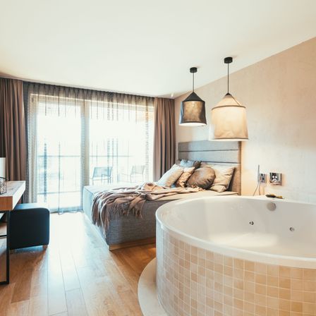 Ein modernes Schlafzimmer mit einem großen Bett, beigefarbenen Wänden und einer großen Badewanne. Es gibt zwei Lampen und einen Schreibtisch mit einem Hocker.