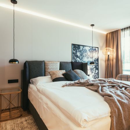 Ein Schlafzimmer mit einem Bett, geschmückt mit verschiedenen Kissen, einer Decke und einer Lampe. Ein Bilderrahmen hängt an der Wand, und ein Fenster mit Vorhängen ist sichtbar.