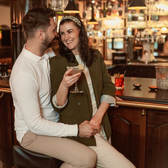 Ein Mann und eine Frau sitzen gemeinsam an einer Bartheke, lächeln und halten einen Cocktail in der Hand. Die Bar ist gut beleuchtet, und hinter ihnen sind Regale mit Flaschen und Gläsern zu sehen.