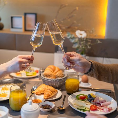 Zwei Hände stoßen mit Champagnerflöten über einem Frühstückstisch an, der mit Croissants, Eiern und Obst gefüllt ist. Dahinter schmücken gerahmte Bilder die Wand.