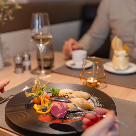 Ein schwarzer Teller mit Gebäck und Sorbet darauf. Es gibt eine Gabel in der Hand. Der Tisch hat eine Blumenvase, ein Weinglas und eine Tasse.