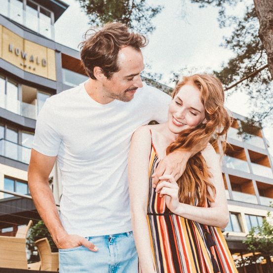 Ein Paar steht lächelnd eng beieinander vor einem Gebäude mit dem Schriftzug 'ROYALD'. Der Mann hat seinen Arm um die Frau gelegt. Hinter ihnen befinden sich Stühle und Bäume.