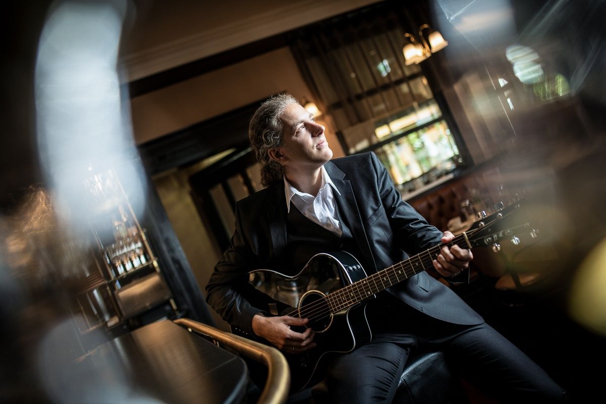 Ein Mann in einem schwarzen Anzug spielt eine Gitarre in einem schwach beleuchteten Raum. Die Gitarre hat einen schwarzen Korpus und goldene Akzente. Die Augen des Mannes sind geschlossen, während er spielt.