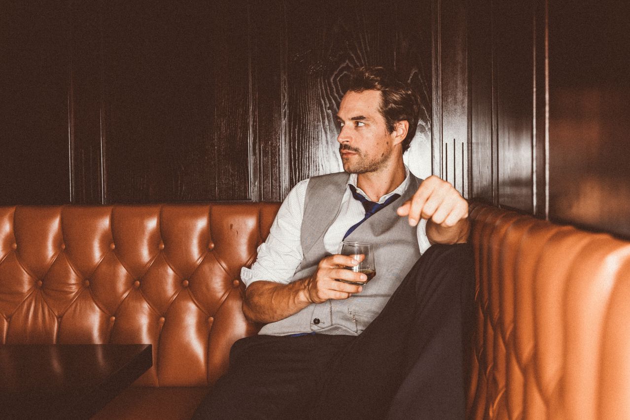 Ein Mann in formeller Kleidung sitzt auf einer braunen Ledercouch, hält ein Glas Whiskey und zeigt mit dem Finger.