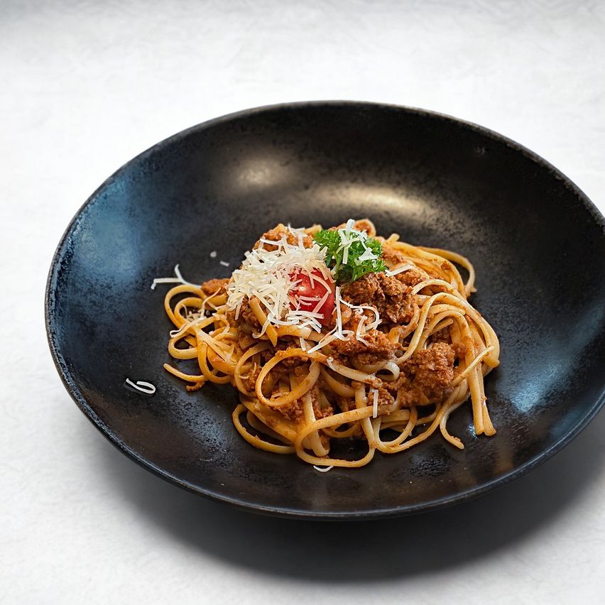 A black bowl with a plate of spaghetti topped with ground beef, grated cheese, and a slice of tomato.