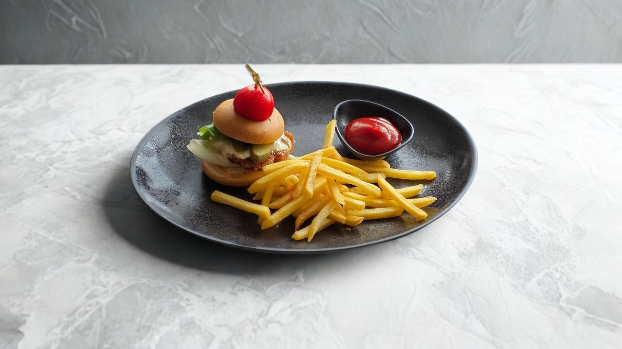 A black plate with a chicken burger topped with a cherry tomato and a side of French fries, accompanied by a small bowl of ketchup.