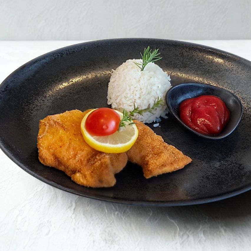 A black plate with fried fish, rice, a lemon slice, a tomato, and a bowl of ketchup.