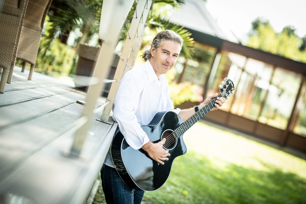 Ein Mann in einem weißen Hemd und Jeans steht auf einer Holzterrasse, lächelt und hält eine schwarze akustische Gitarre.