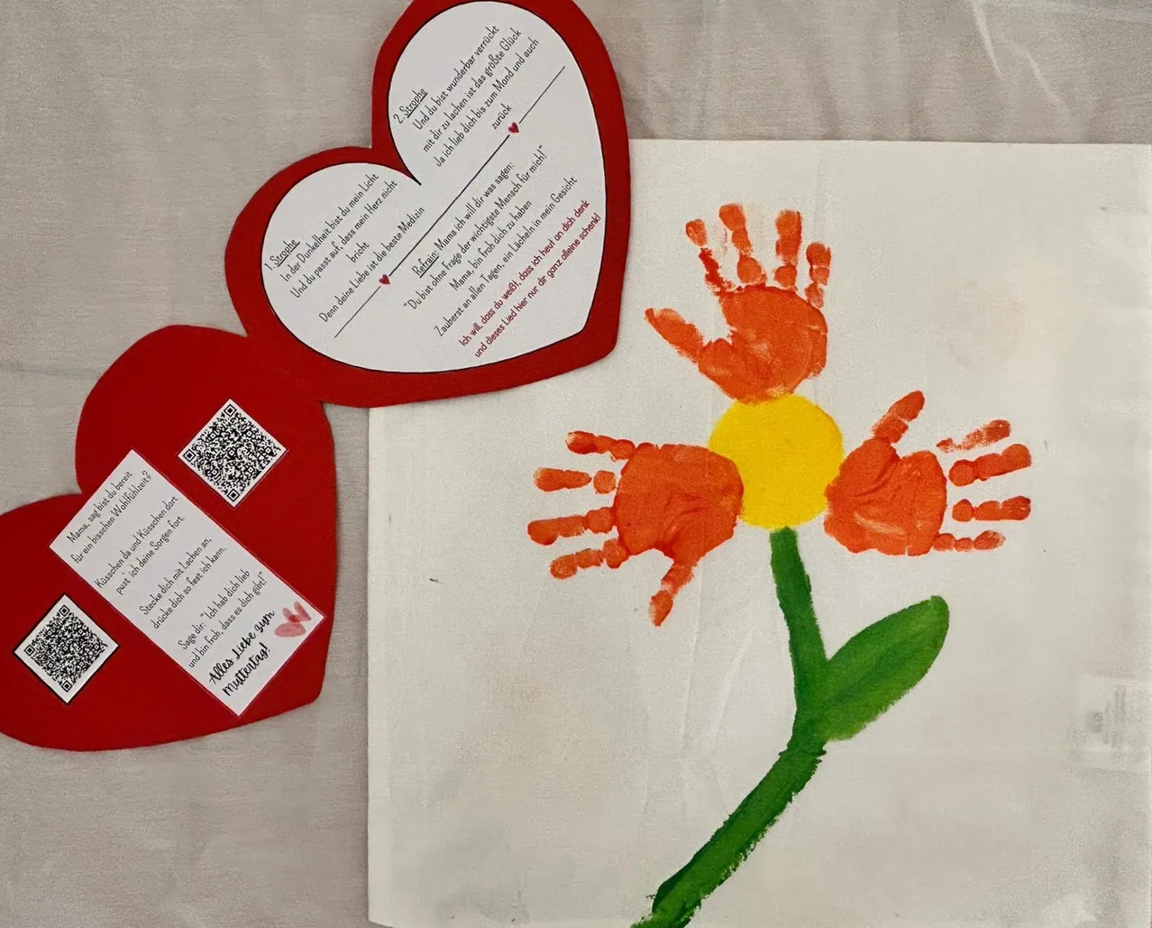 Ein herzförmiges Papierbastelwerk mit einer Blume und Handabdrücken. Ein Gedicht ist auf dem Herz geschrieben, und ein QR-Code befindet sich auf der linken Seite.