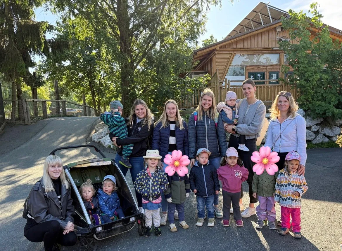 Eine Gruppe von Frauen und Kindern steht vor einem Holzgebäude, einige halten rosa Luftballons. Einige tragen Babys und andere halten die Hände von Kindern.