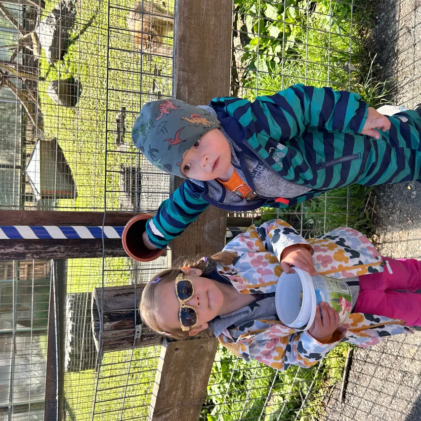 Zwei Kinder stehen neben einem Zaun in einem Zoo. Ein Kind hält einen Eimer, das andere einen kleinen Topf. Sie schauen zur Kamera hoch und tragen warme Kleidung.