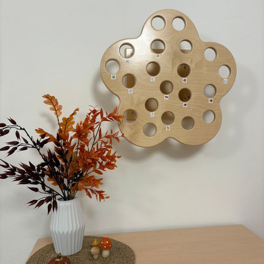 A wooden flower-shaped object with holes and small tags hangs on a white wall, next to a vase with autumn leaves.
