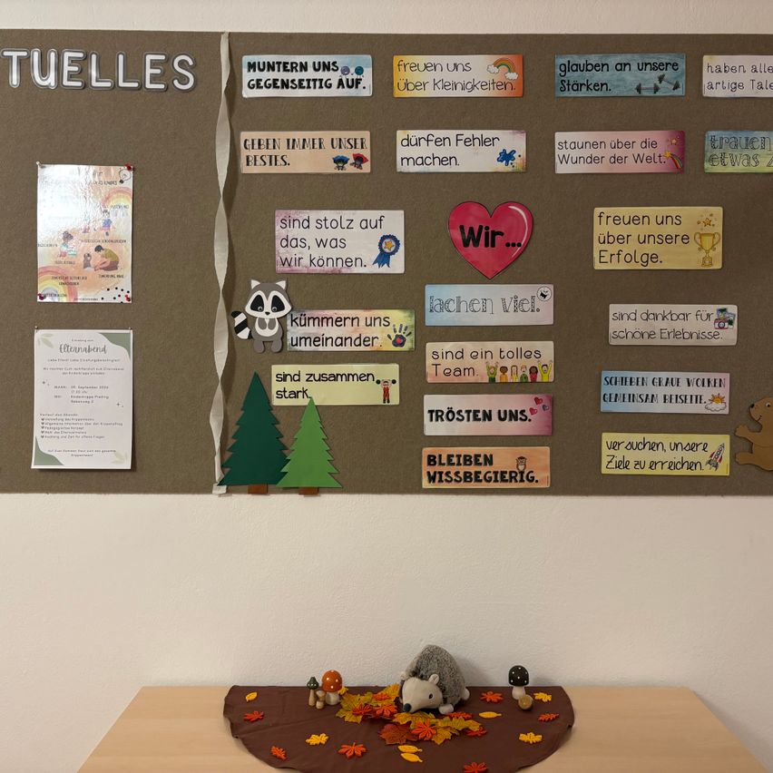 A bulletin board with various colorful cards and messages, including a red heart, a raccoon, and a tree. Below the board, there is a table with autumn leaves and small animal figurines.
