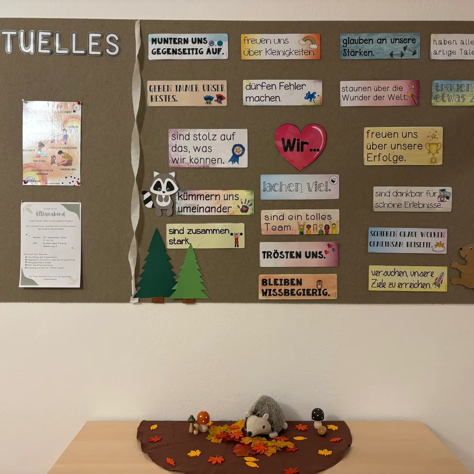 Ein Bulletinboard mit verschiedenen bunten Karten und Nachrichten, einschließlich eines roten Herzens, eines Waschbären und eines Baumes. Unter dem Board befindet sich ein Tisch mit Herbstblättern und kleinen Tierfiguren.