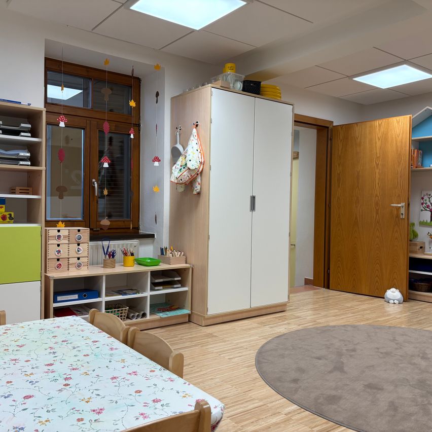 A well-organized playroom with wooden floors, a table covered in a floral tablecloth, a cabinet with shelves, and a rug. The room is decorated with colorful mushroom-themed decorations.