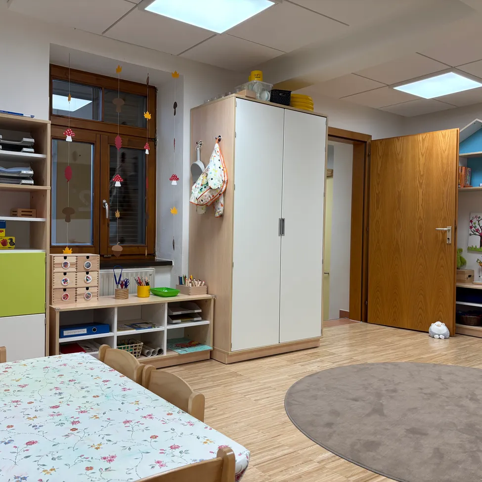 Ein gut organisiertes Spielzimmer mit Holzboden, einem mit einem Blumentischtuch bedeckten Tisch, einem Schrank mit Regalen und einem Teppich. Der Raum ist mit farbenfrohen, pilzthematischen Dekorationen geschmückt.