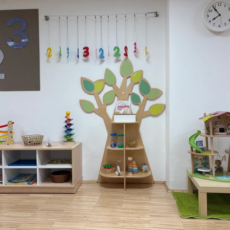 Ein Kinderspielzimmer mit einem hölzernen Baum, Regalen, Spielzeug und einer Wand mit Zahlen und einer Uhr.
