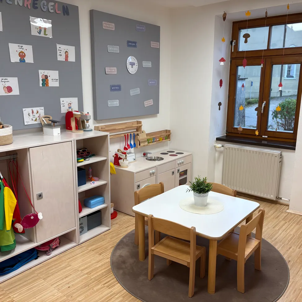 Ein kleiner, gut organisierter Spielraum mit einem weißen Tisch und Stühlen, einer kleinen Küchenzeile und einer Wand mit verschiedenen Postern.