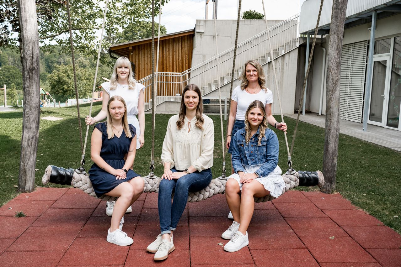 Five women are sitting on a swing, smiling and posing for a photo. They are dressed casually and are sitting on a red platform. Behind them is a building with stairs and a wooden wall.