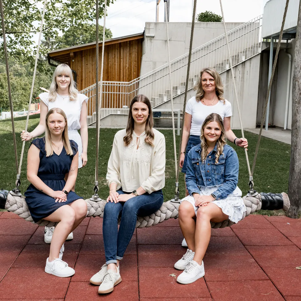 Fünf Frauen sitzen auf einer Schaukel und posieren für ein Foto. Sie sind lässig gekleidet und sitzen auf einer roten Plattform. Hinter ihnen befindet sich ein Gebäude mit Treppen und einer Holzwand.