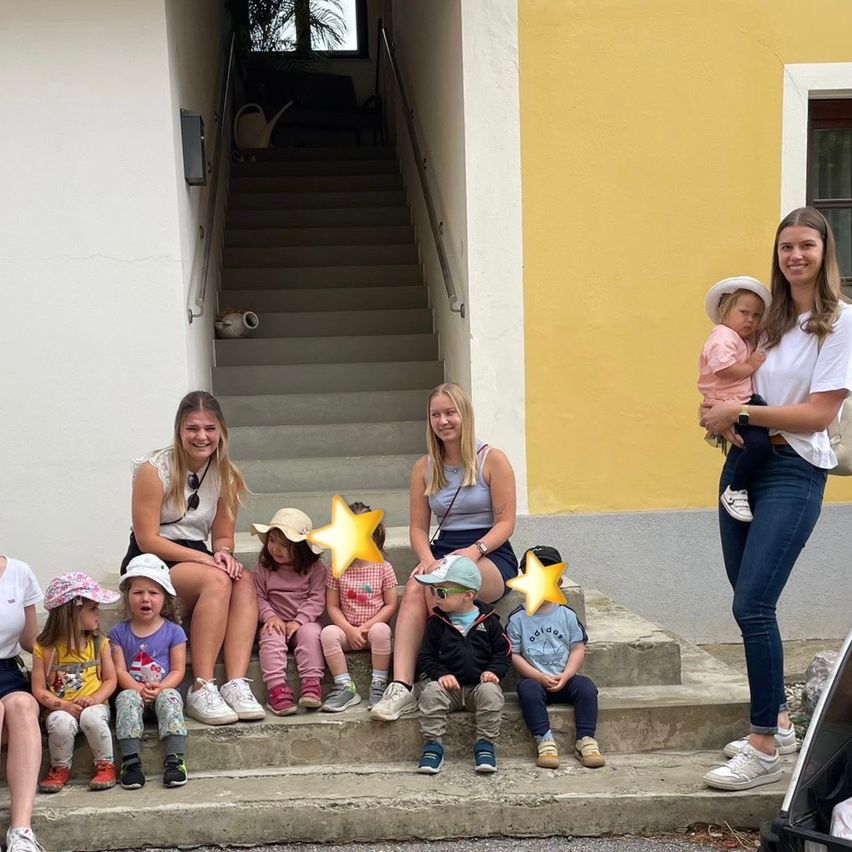 Eine Gruppe von Kindern und Erwachsenen sitzt auf den Stufen eines Gebäudes. Sie alle lächeln und schauen nach vorne. Das Gebäude hat eine gelbe Wand.