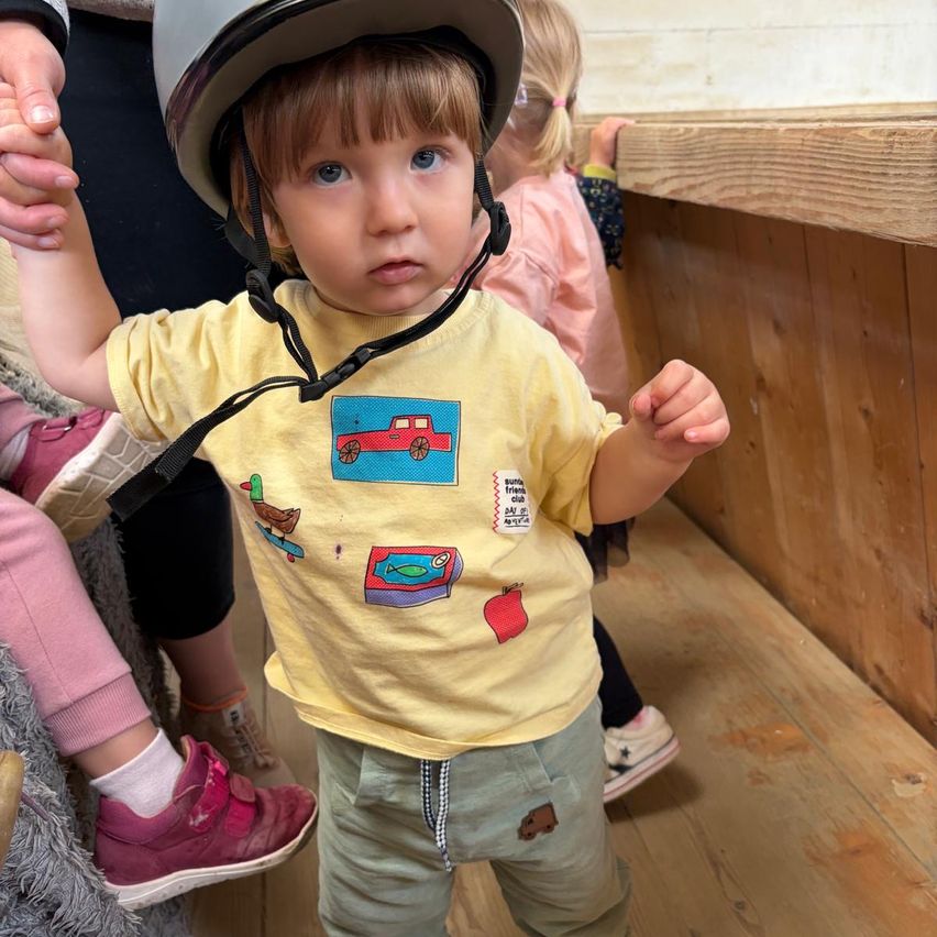 Ein Kleinkind trägt ein gelbes T-Shirt mit verschiedenen Cartoon-Bildern und einem Helm und steht auf einem Holzboden. Dahinter hält eine Person seine Hand. Ein weiteres Kind steht in der Nähe, und im Hintergrund sind Holzstrukturen zu sehen.
