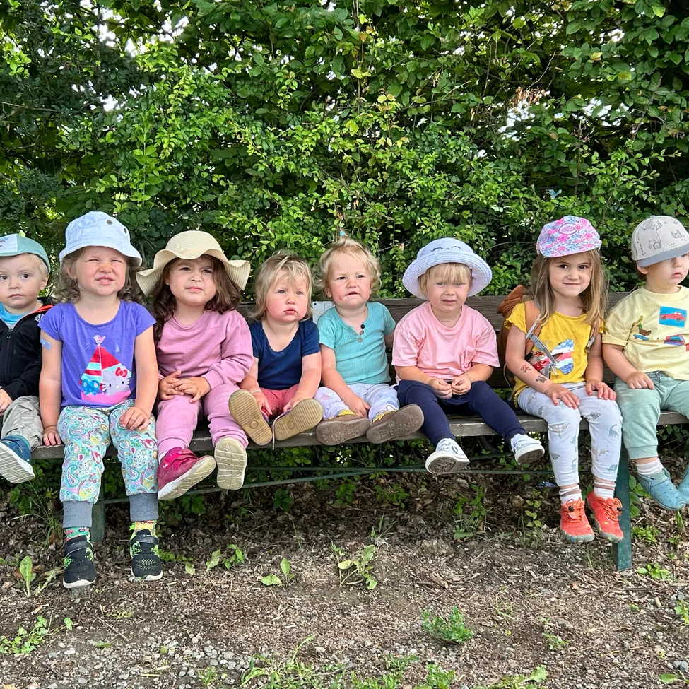 Eine Gruppe von Kindern sitzt auf einer Bank und trägt Hüte und Schuhe. Sie schauen in verschiedene Richtungen. Sie sind von Grün umgeben.