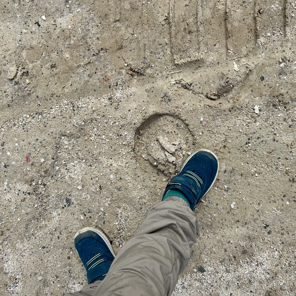 Eine Person steht auf sandigem Boden und trägt blaue Schuhe. Fußabdrücke sind sichtbar, einer bildet ein Herz.