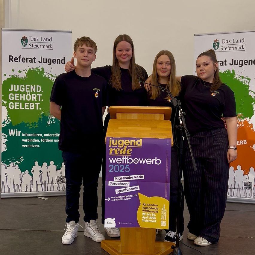 Vier Menschen in schwarzen T-Shirts stehen hinter einem Podium mit der Aufschrift Jugend Rede Wettbewerb 2025. Sie posieren für ein Foto. Dahinter befinden sich zwei Plakate. Das linke Plakat hat die Aufschrift Referat Jugend. Das rechte Plakat hat die Aufschrift Jugend. Gehört. Gelebt.