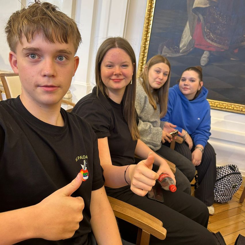 Vier Menschen sitzen auf Stühlen in einem Raum. Der Junge links gibt einen Daumen hoch. Das Mädchen neben ihm hält eine Flasche Coca-Cola. Ein Gemälde befindet sich an der Wand hinter ihnen.
