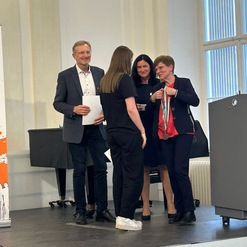 Drei Frauen und ein Mann stehen auf einer Bühne. Der Mann hält ein Papier und die Frau neben ihm eine Medaille. Die Frau auf der rechten Seite hält ein Mikrofon. Hinter ihnen steht ein Klavier und ein Heizkörper.