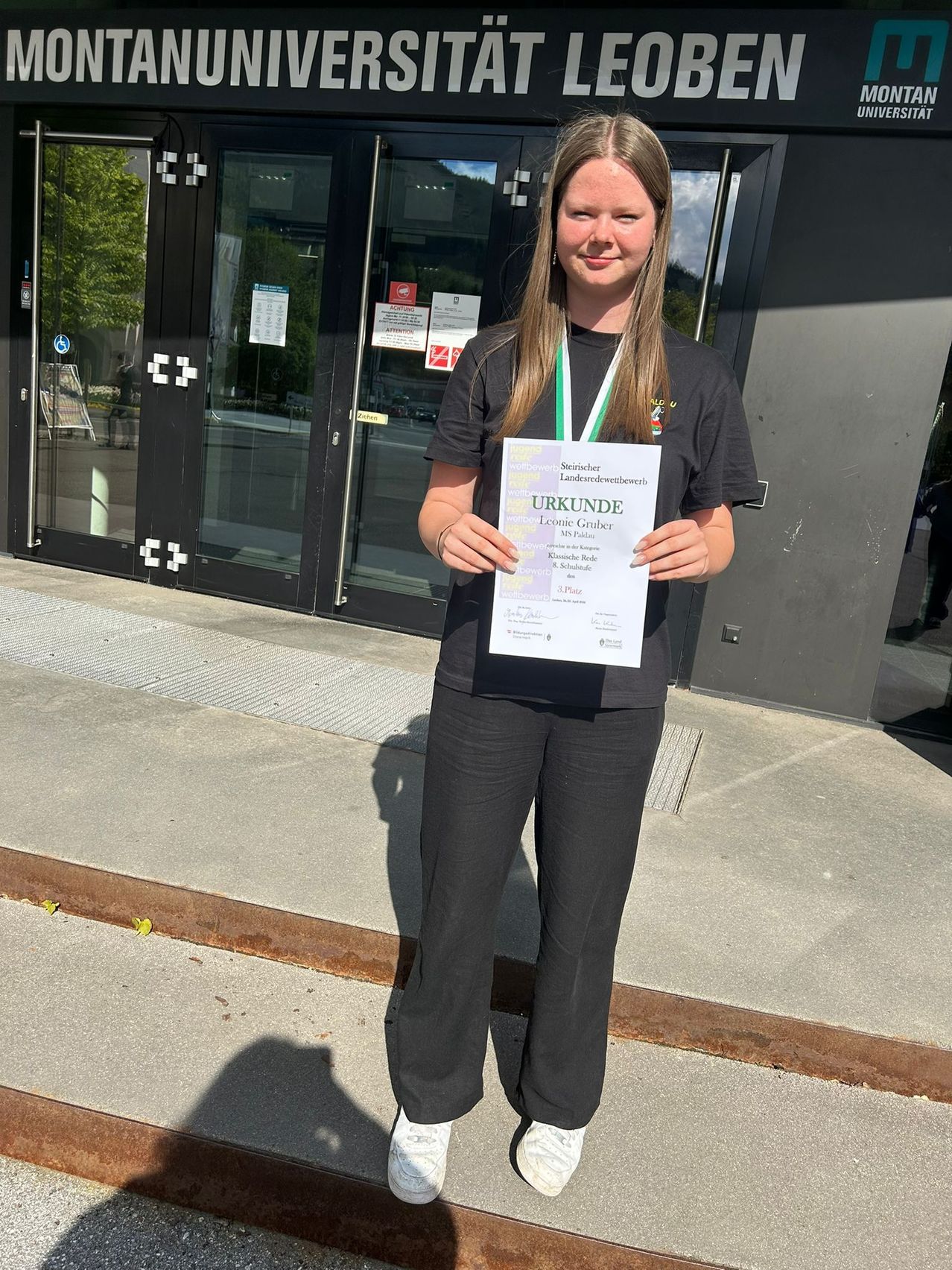 Eine Frau in einem schwarzen Shirt und Hose posiert mit einem Zertifikat vor einer Glastür eines Gebäudes.