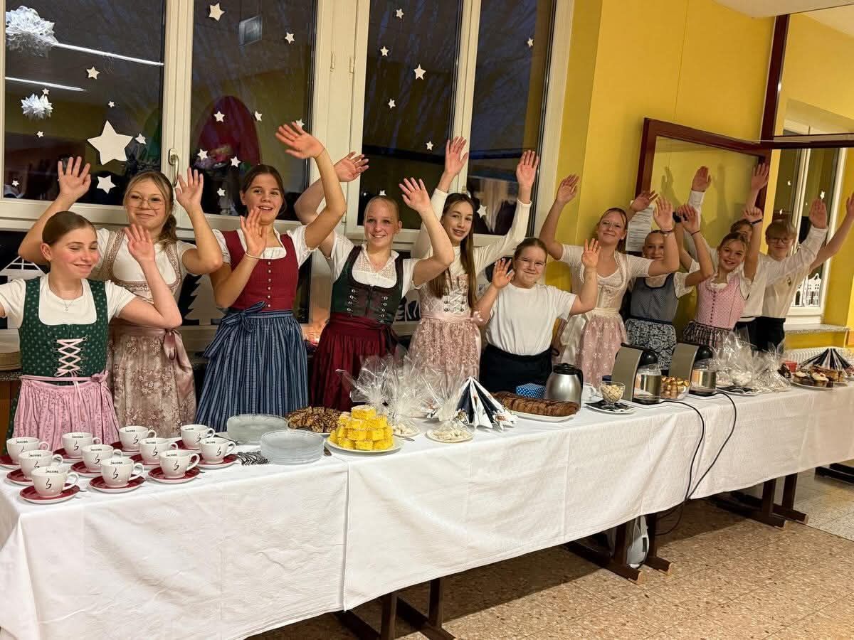 Eine Gruppe von Frauen in traditioneller Kleidung hebt ihre Hände vor einem Tisch mit verschiedenen Lebensmitteln. Der Tisch ist mit einem weißen Tuch bedeckt.