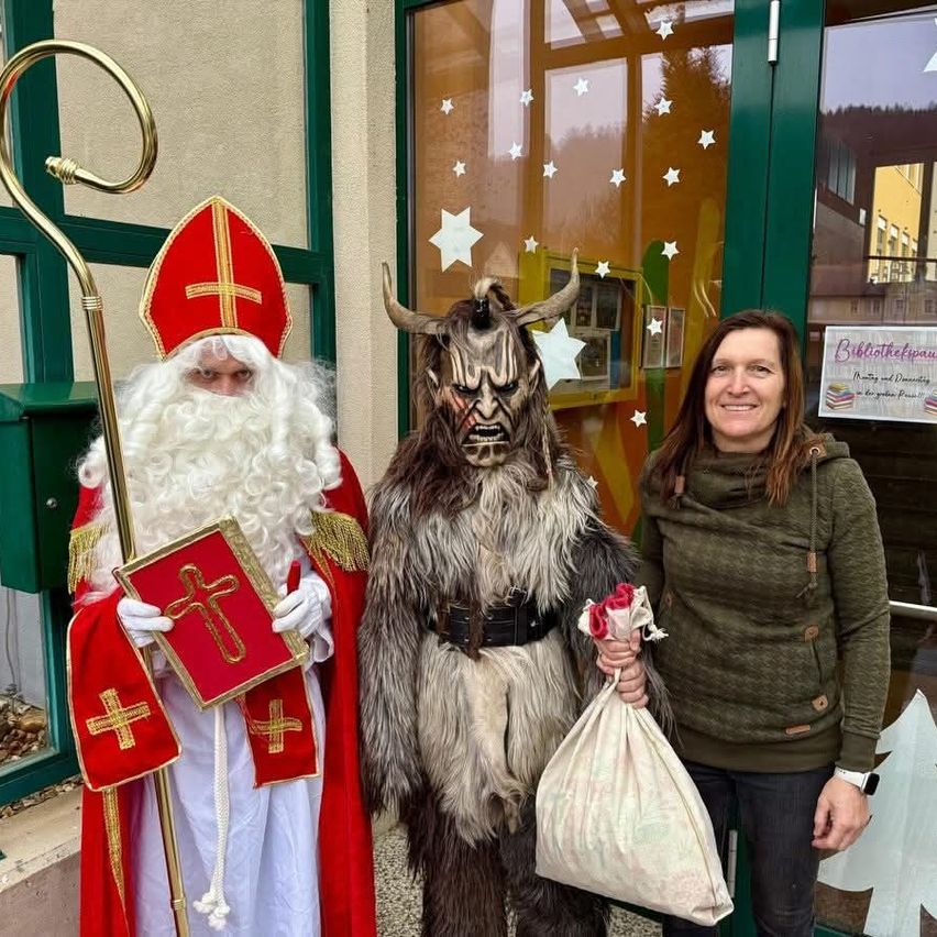 Eine Frau in einem grünen Hoodie steht zwischen zwei Männern, einem in einem Weihnachtsmann-Kostüm und dem anderen in einem Krampus-Kostüm. Die Frau hält einen Beutel und lächelt in die Kamera.