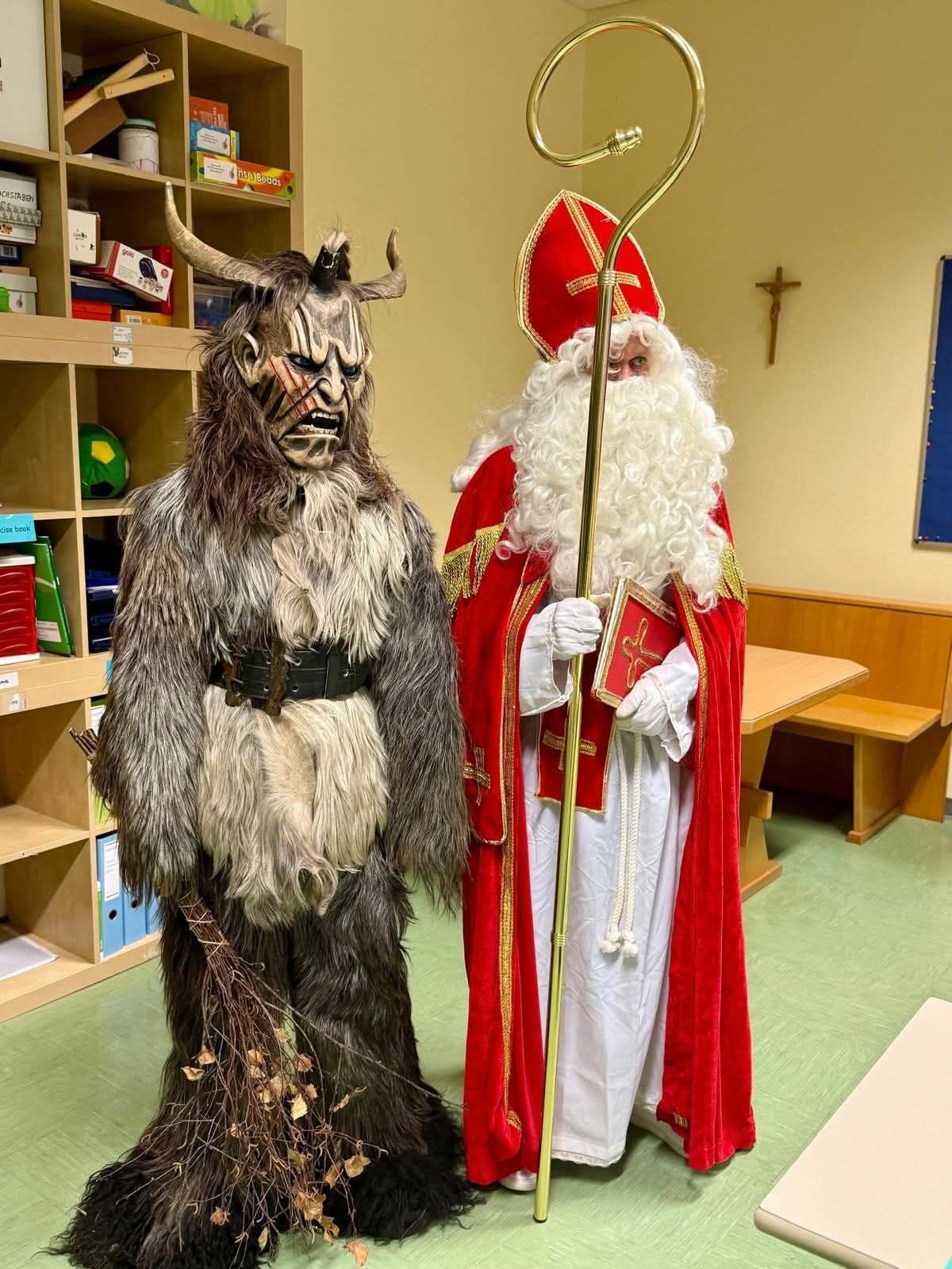 Zwei Personen in Kostümen stehen nebeneinander in einem Raum. Einer ist als Dämon mit Hörnern und einem Gürtel gekleidet, während der andere als Weihnachtsmann mit einem Stab gekleidet ist. Hinter ihnen steht ein Regal mit verschiedenen Gegenständen.