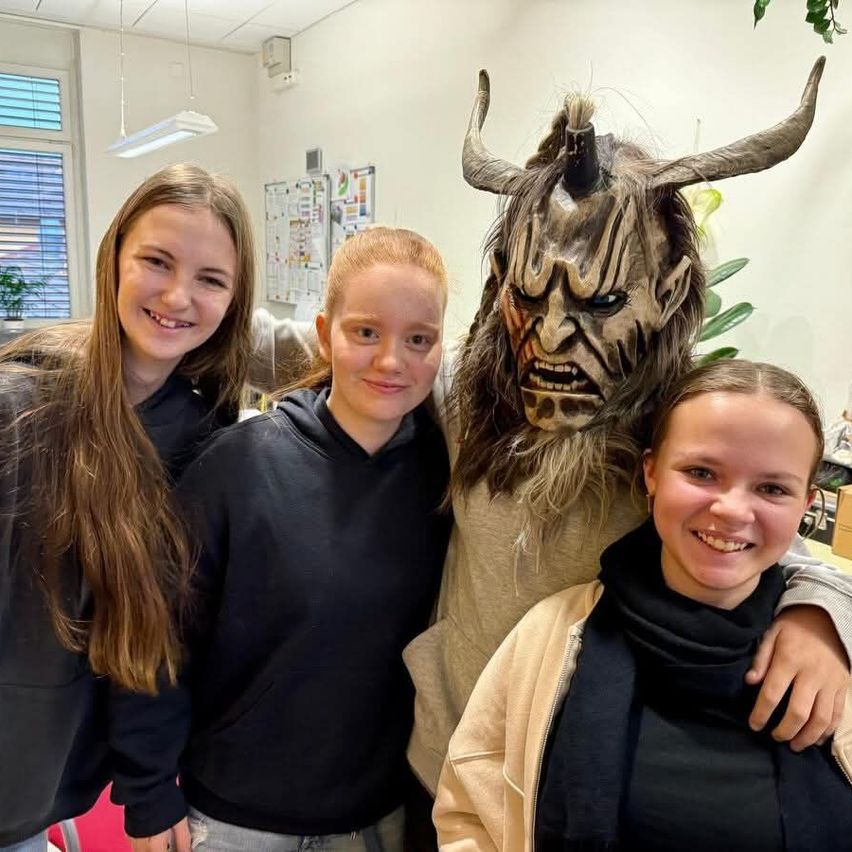 Drei Mädchen und eine Person mit Hornmaske lächeln für ein Foto in einem Raum mit weißen Wänden und einem Fenster.