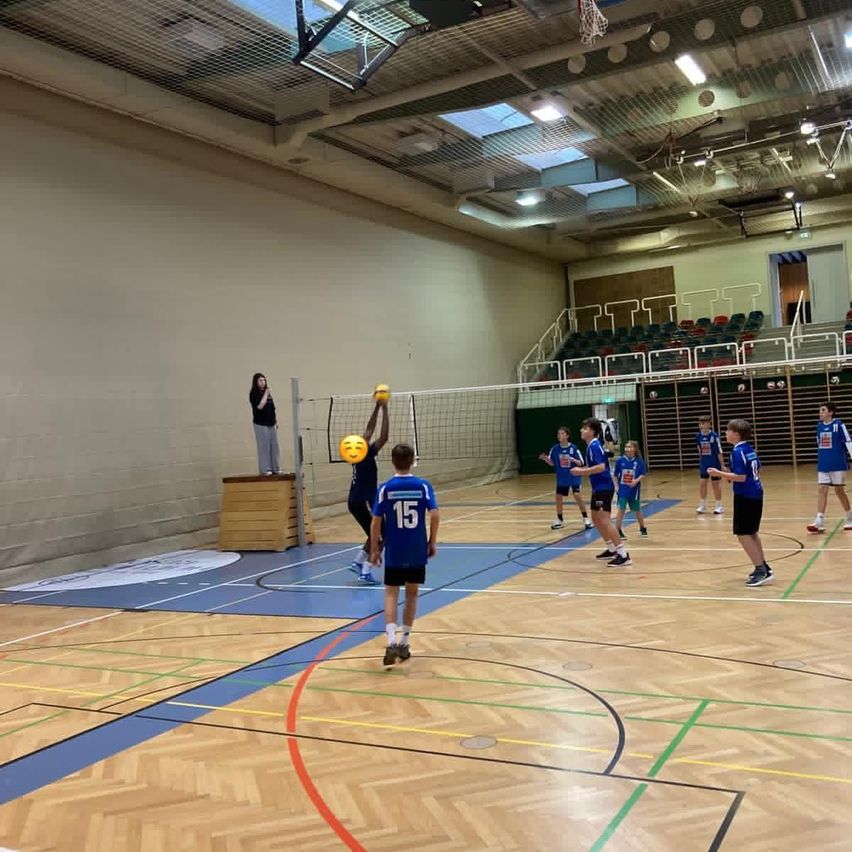 Eine Gruppe junger Jungen spielt Volleyball in einer Turnhalle. Ein Junge springt, um den Ball zu schlagen, während ein anderer Junge hinter dem Netz steht.