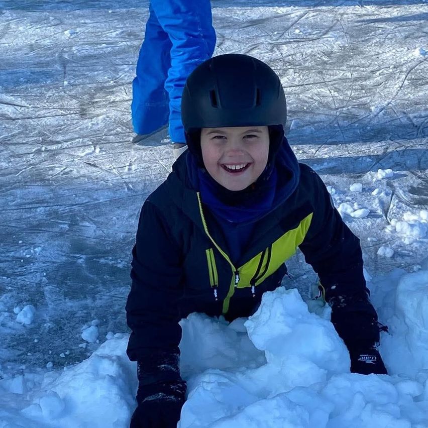 Ein junger Junge in einem schwarzen Helm und einer Jacke lächelt, während er auf einer Eisbahn durch den Schnee kriecht.
