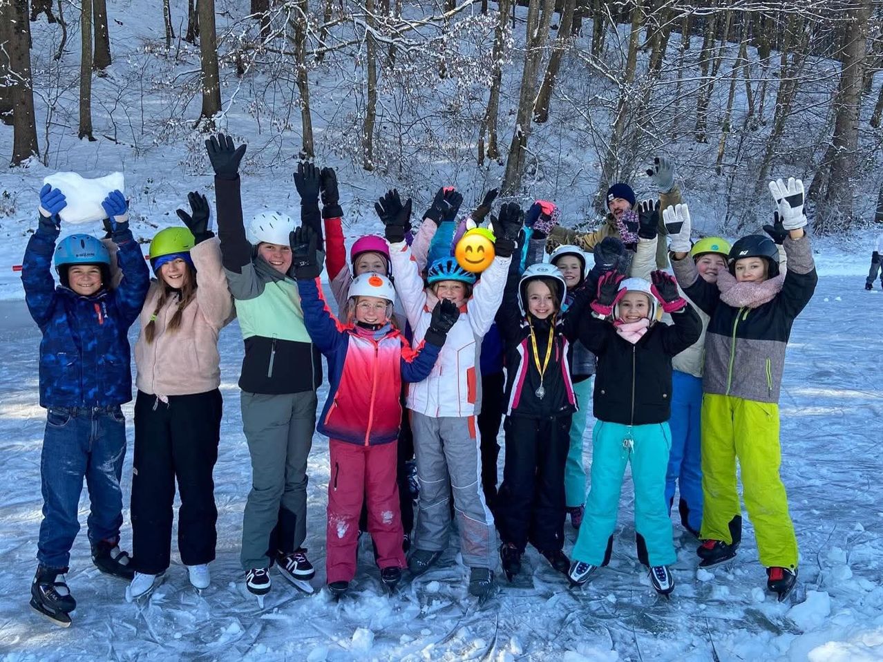 Eine Gruppe von Kindern und Erwachsenen steht auf Eis, trägt Winterkleidung, Handschuhe und Helme. Sie lächeln und posieren für ein Foto in einem verschneiten Wald.