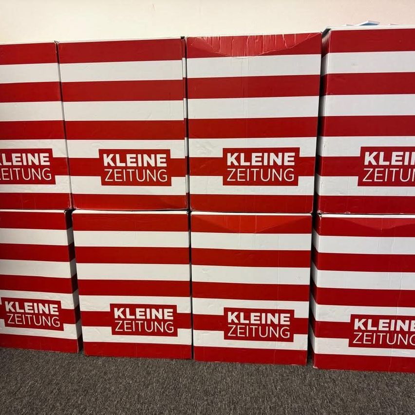 Several red and white boxes are stacked, labeled 'Kleine Zeitung.' They are arranged neatly on a gray floor.