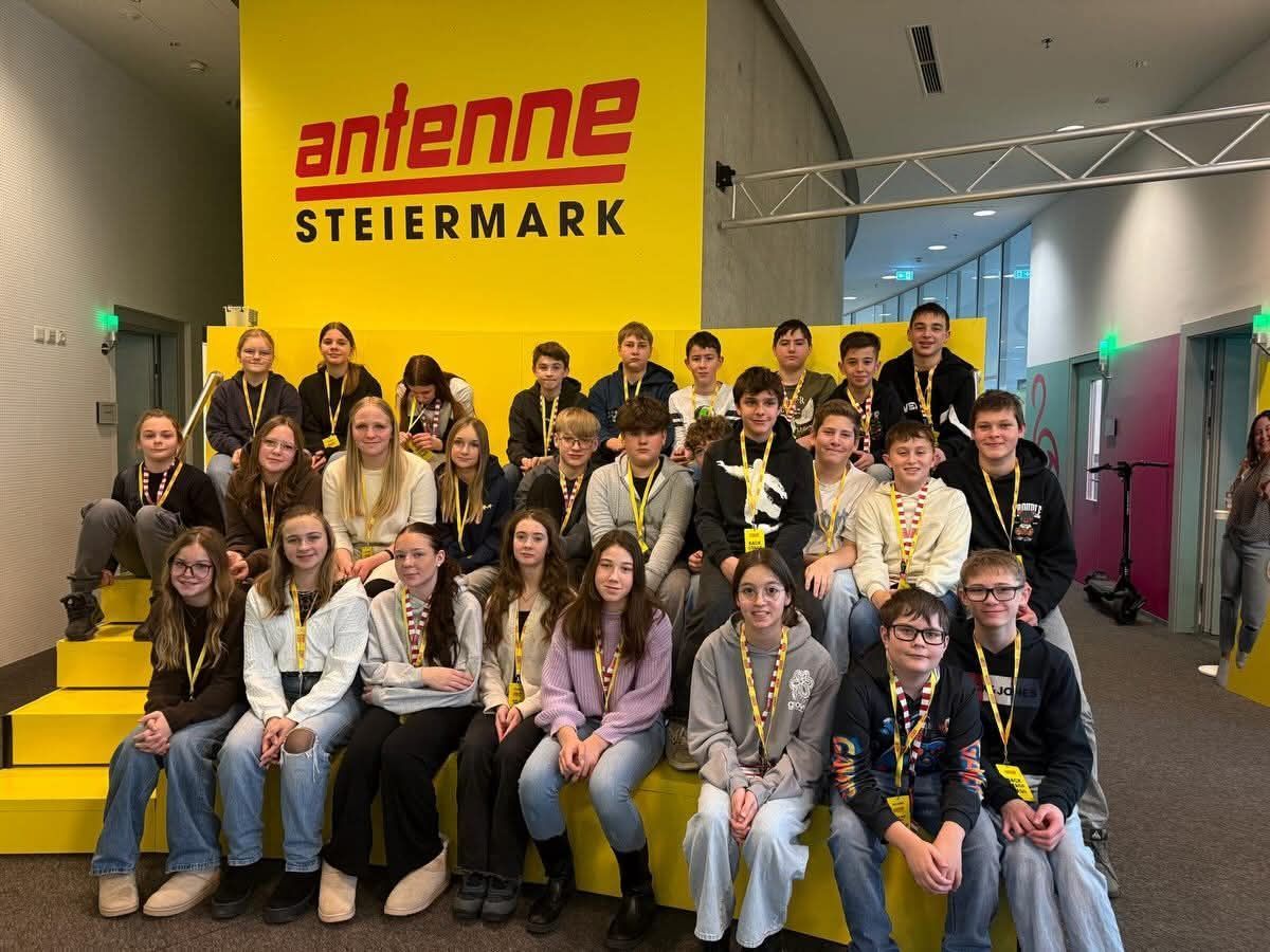 A group of young people are posing for a photo in front of a yellow banner that reads 'Antenne Steiermark'. They are wearing IDs and some are wearing glasses. They are sitting on a yellow bench.