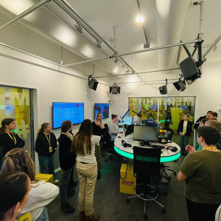 In a studio, a group of people are gathered around a round desk with monitors and a computer. A woman is speaking into a microphone, and another woman is listening.