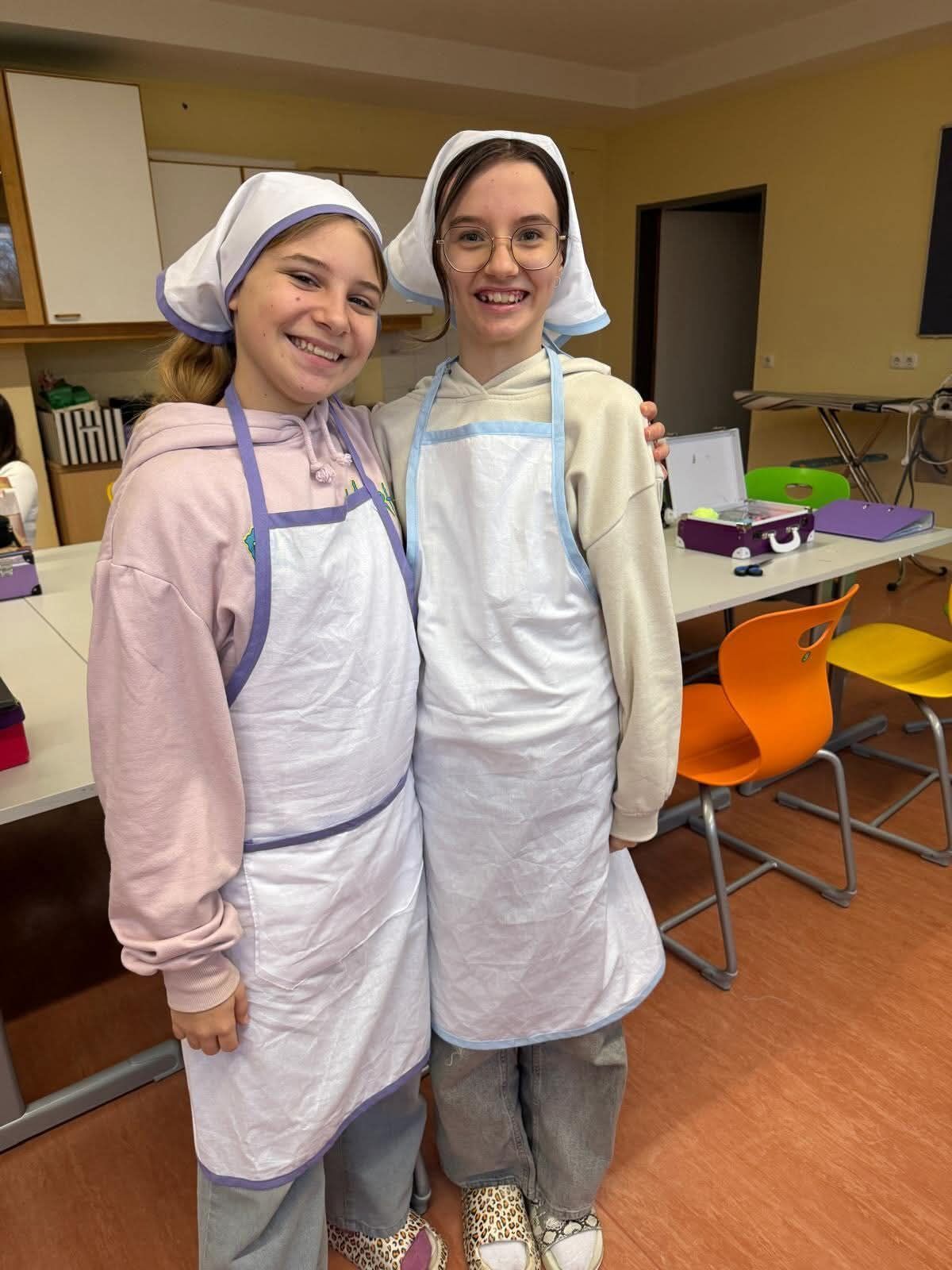 Two young girls in aprons and chef hats are smiling in a classroom with a table and chairs in the background.