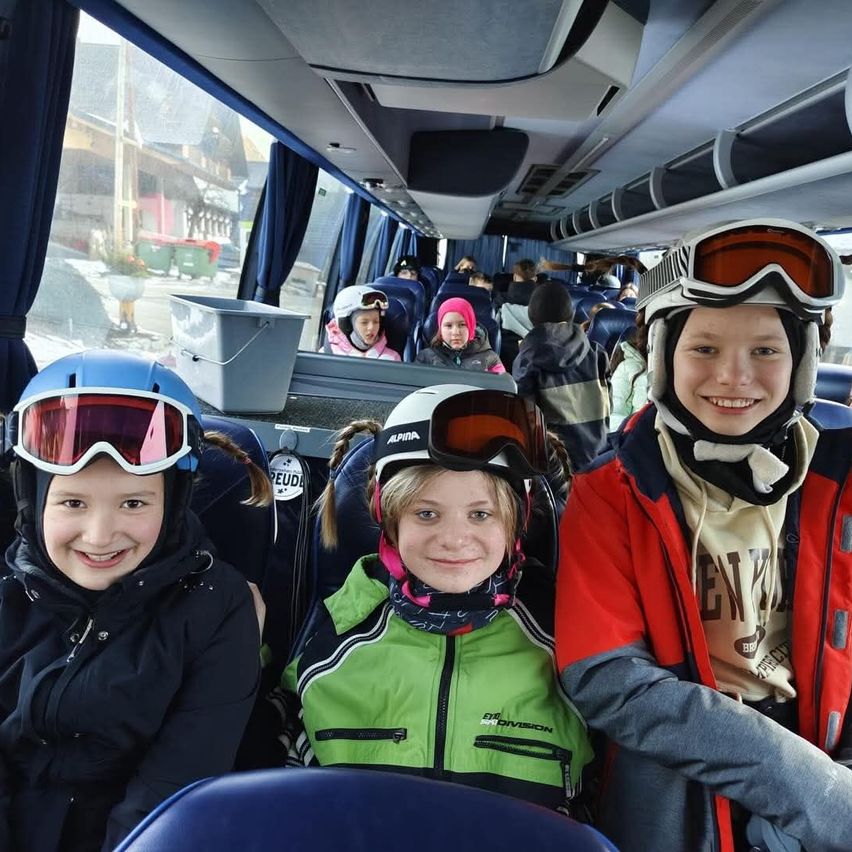 Drei Kinder in Skiausrüstung lächeln für ein Foto im Bus, mit anderen Passagieren und Außenszenen, die durch die Fenster sichtbar sind.