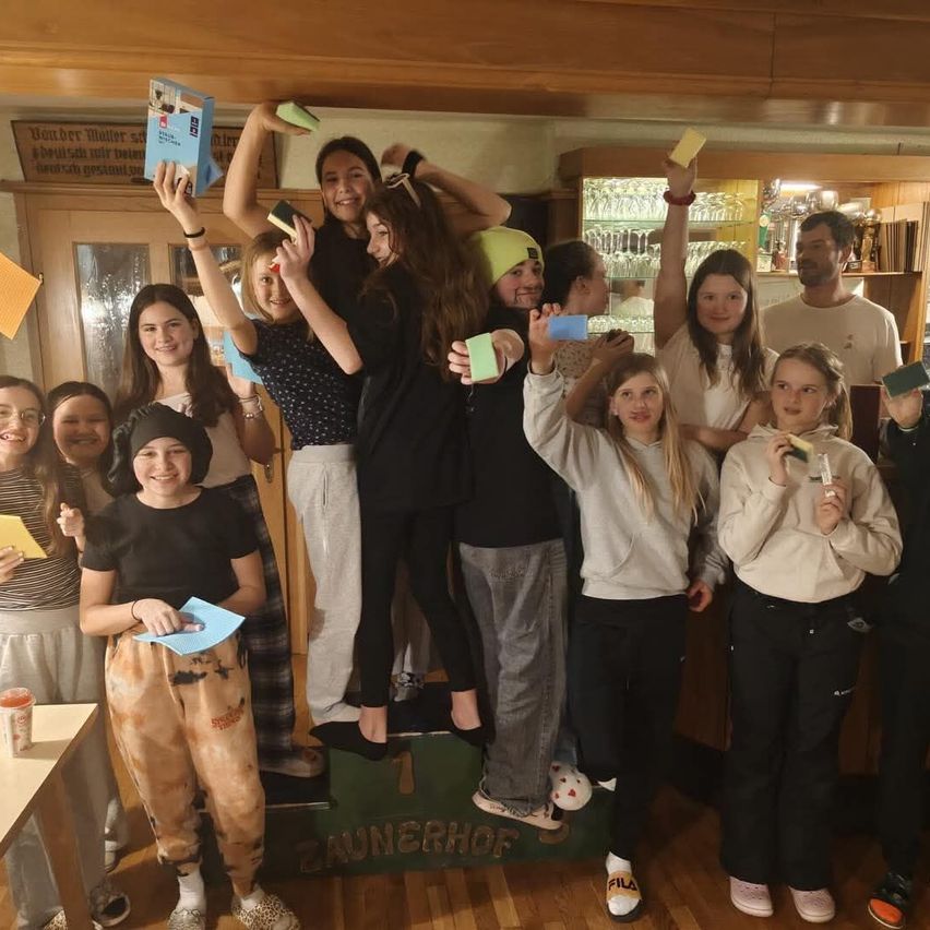 Eine Gruppe junger Mädchen und Jungen posiert für ein Foto in einem Raum. Einige halten Papiere und Reinigungsmittel. Ein Holzregal mit Gläsern ist im Hintergrund.