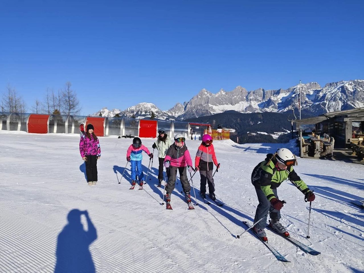 Eine Gruppe von Kindern schneit mit Helmen und Skistöcken einen schneebedeckten Berghang hinunter, unter einem klaren blauen Himmel mit Bergen im Hintergrund.