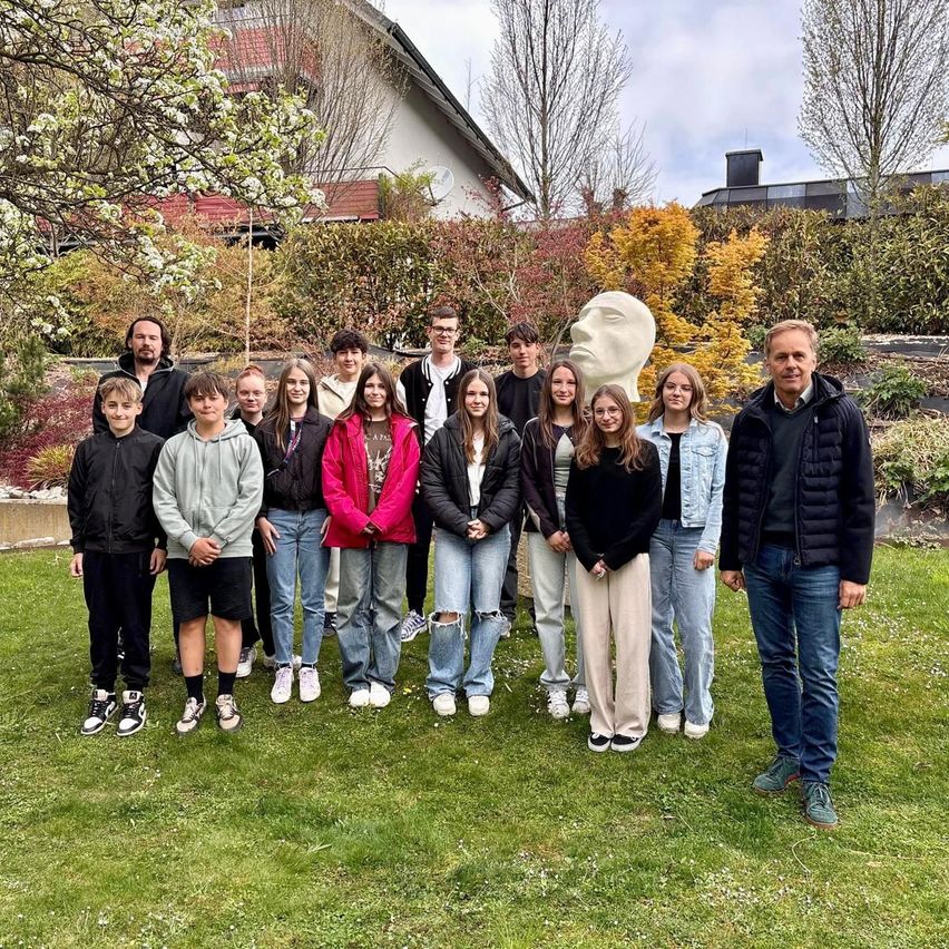 A group of young people and two men pose in front of a garden with a sculpture and a house in the background.
