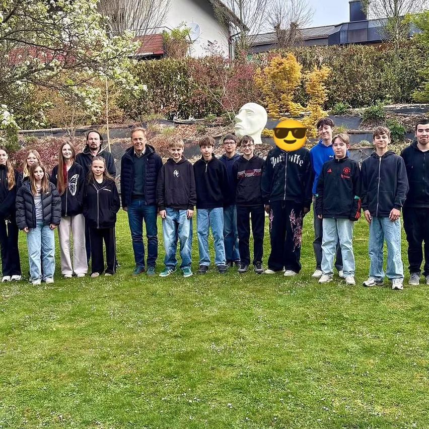 A group of people stands on a lawn, some in jackets and sneakers, one in sunglasses. Trees and plants are behind them, and a statue in the middle.